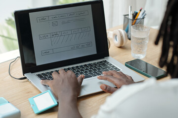 Person engrossed in designing digital user interface layout on laptop. Various devices like smartphone and headphones present on desk suggesting tech-focused task