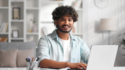 Portrait of a young Indian man working at a computer in his office, turns his head to look at the camera and smiles sincerely. Close up.