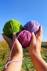 Green, purple, and red wool threads held high against a bright blue sky on a sunny day, celebrating the joy of knitting as a favorite hobby.