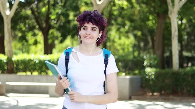 Video of a young student with purple curly hair walking through a tree-lined city area with a backpack and earphones, as the camera pans to the side