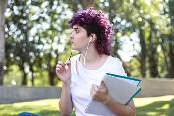 Young nonbinary person with purple curly hair wearing earphones and thinking ideas while holding a...