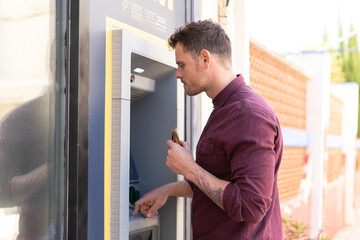 Fototapeta premium Young caucasian man at outdoors using an ATM