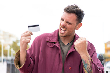 Young caucasian man holding a credit card at outdoors celebrating a victory