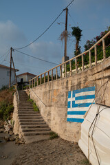 Greek flag painted on a cement wall with stone staircase and metal railings heading from the sandy...