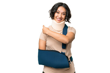 Young argentinian woman wearing neck brace and sling over isolated background pointing back
