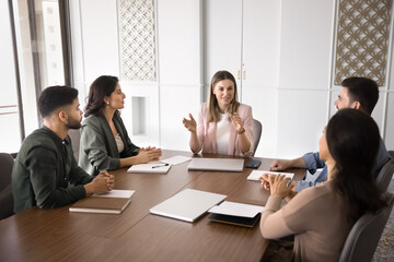 Fototapeta premium Positive young project leader woman holding meeting with team, talking to employees, coworkers at large table, offering marketing strategy, teamwork plan, ideas for brainstorming