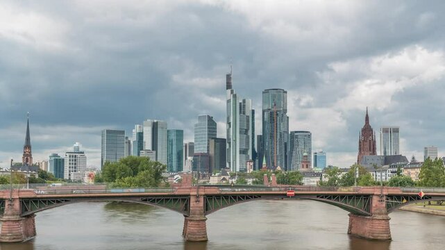 Aerial hyperlapse of Frankfurt skyline with Main River and Ignatz Bubis Bridge. Skyscrapers and church spires reflected in the water beneath dramatic clouds timelapse. Frankfurt, Germany