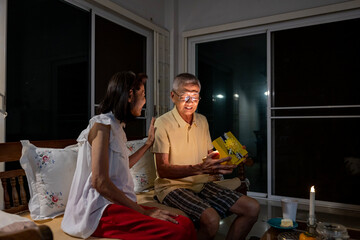 Senior asian couple is happily celebrating birthday together at home while exchanging gift box present for love, relationship, bonding, longevity and marriage