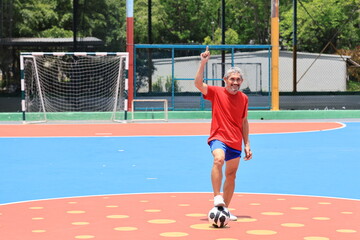 Fototapeta premium Senior Asian man in retirement age playing soccer football at outdoor court for healthy and fun activity to exercise and workout against aging illness