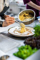 A chef’s hand plating a gourmet dish with a spoon in a professional kitchen. The white plate features a delicate arrangement of food with sauce, showcasing culinary artistry and attention to detail