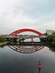 Fototapeta premium Traffic bridge over river in Ho Chi Minh City, Vietnam from aerial view featuring two large red steel arches and a wide, ten lane highway for heavy vehicles. Low angle with reflection on water