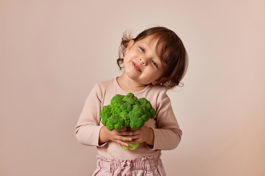 smiling child girl holding fresh green raw broccoli on beige background. healthy baby food. kid loves vegetables