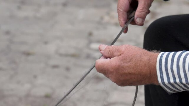 Sailor in striped shirt repairs an old net with knife before fishing. Fisherman holds knife in hand and rope in other, cutting the line. Hobbies and fishing. Close up of elderly mans hands