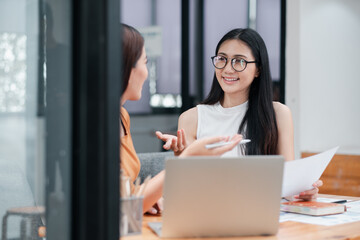Two women discussing work at a modern office, using a laptop and documents, showcasing teamwork and...