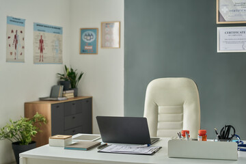 Contemporary medical office featuring an organized desk, laptop, and certificates on walls. Plants and storage cabinet add warmth to professional space