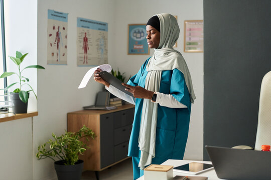 Muslim African American female doctor standing in medical office, holding patient files and reviewing data. Room decorated with educational charts and plants