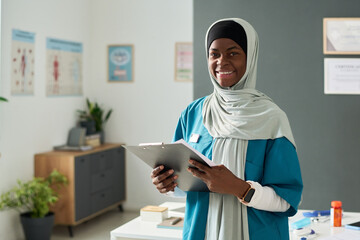 Smiling Muslim female healthcare professional standing in office wearing traditional hijab and...
