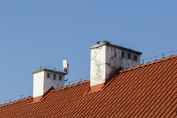 Old orange ceramic roof tiles. Tiled roof texture. Blue sky over the house. Chimney wooden vent structure at the rooftop's peak. Lightning rod running along the roof. Internet antenna on the roof.