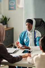 Muslim medical professional wearing scrubs and headscarf explaining medicine to patients in office...
