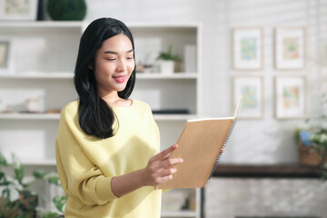 Smiling asian woman happily looks at a note book with names of customers ordering online in preparation for delivery.