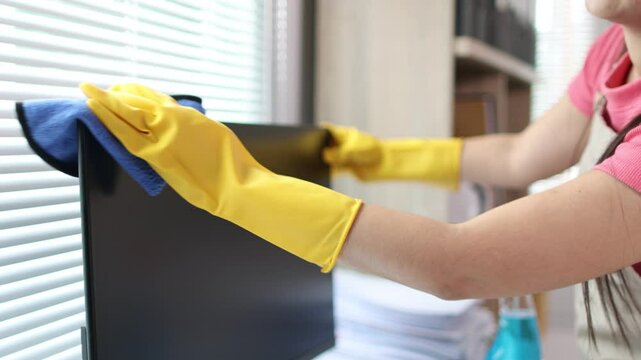 Young Asian cleaning woman uses a cloth to wipe clean a computer and screen using cleaning solution in her home office.
