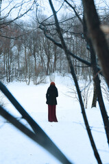 A gothic style, medieval scene. A girl in a long burgundy dress and a black coat goes into the winter forest.