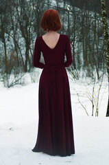 A red-haired girl in a burgundy long dress stands with her back to him. A woman in a gloomy mysterious forest. The Gothic atmosphere.