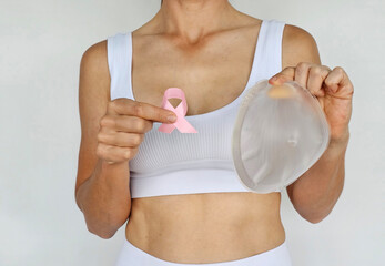 A woman holds a silicone breast implant and a pink ribbon to symbolize breast cancer awareness