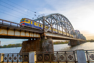 Fototapeta premium A modern electric passenger train crosses the railway bridge (Dzelzcea tilts) over the Daugava River. Taking passengers to the scenic Jurmala region of Latvia.