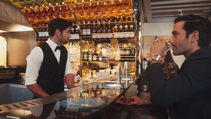 Focusing smiling smart bartender serving gin fizz or tonic soda at luxurious bar, engaging lively conversation with VIP customer couple at back view in atmosphere at lighting warm nightclub. Vinosity.