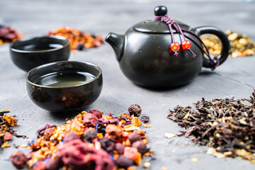 A black teapot and cups on a gray background with scattered tea of different varieties.