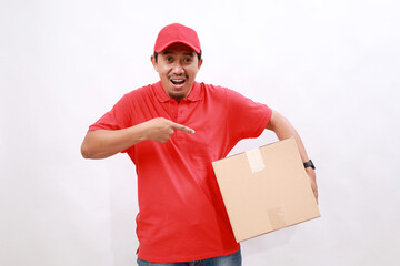 Happy asian delivery courier in red t-shirt and cap carrying a box package while pointing on it. isolated over white background