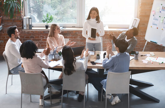 Asian female worker making presentation at business meeting, using digital tablet, multiracial team greeting coworker with applause