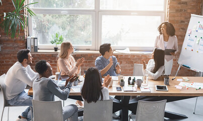 Black creative lady speeking in front of young business team in office, colleagues cheering woman...