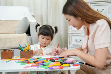 Female Asian teacher assisting focused young girl with colorful pens and blocks in creative activity, encouraging learning through art, strengthening early educational foundation in cozy home setting.