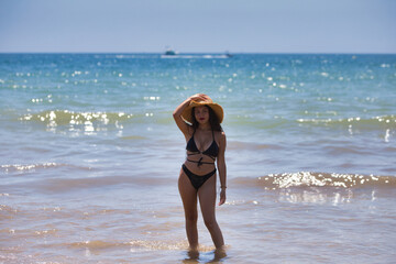 Young and beautiful brunette woman with brown eyes is on the beach in swimsuit and puts a straw hat...