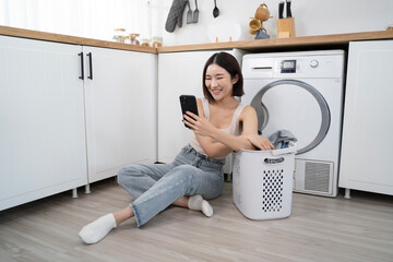 A young Asian woman sits on the floor next to a laundry basket, casually using her phone during her laundry routine in a modern, cozy home setting in relaxation and leisure time with household chores