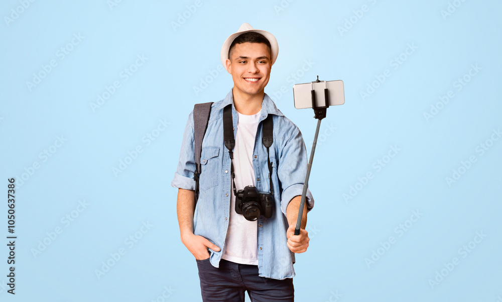 Wall mural positive guy tourist taking photos with smartphone and selfie stick, orange background