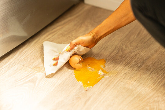 A person is carefully cleaning up a significant egg spill on the floor using a paper towel to wipe it away