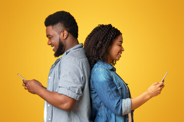 Social Network Addiction. Black Millennial Couple Using Smartphones, Standing Back To Back And Ignoring Each Other, Yellow Background