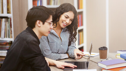 Fototapeta premium Doing Homework Together. Two multiracial students using laptop sitting at desk in modern library, copyspace