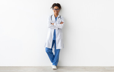Friendly female black doctor in glasses posing with folded arms looking at camera standing leaning...