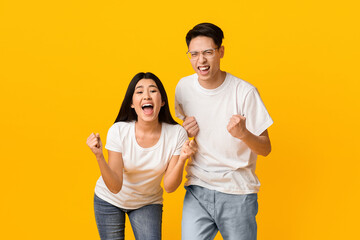 Obraz premium Asian partners rejoicing success, clenching fists and exclaiming with triumph, standing next to each other on yellow background