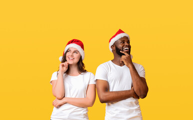 Obraz premium Making holiday plans. Pensive interracial couple wearing santa hats and posing with thoughtful face expression over yellow background, panorama