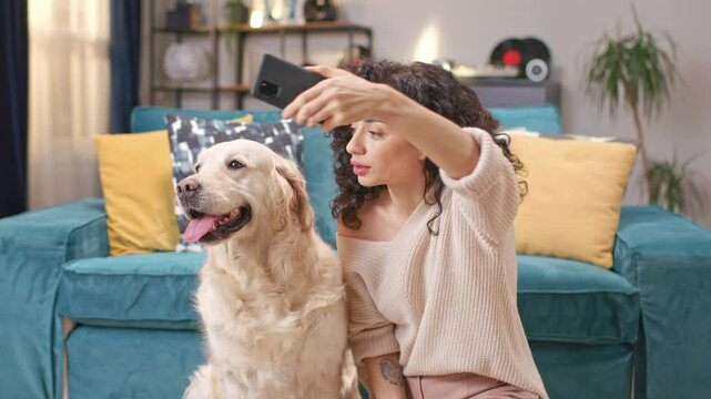Portrait of beautiful smiling young woman taking selfie or making video call with her smartphone while sitting at floor with her cute fuzzy dog. Cozy moments, happiness with lovely pet.