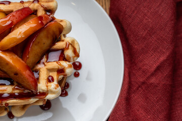 Belgian waffles with apples stewed in wine sauce on a white plate on a wooden background with a burgundy napkin. Place for text