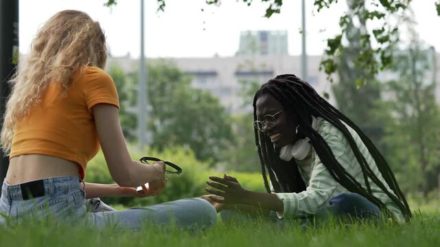 Two young women are sitting on the grass in a park, playing a hand-clapping game. They are both smiling and laughing, enjoying their time together
