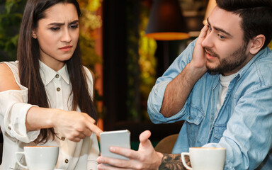 Infidelity Concept. Jealous Girlfriend Pointing Finger At Cheating Boyfriend's Phone Demanding Explanation Sitting In Cafeteria. Selective Focus