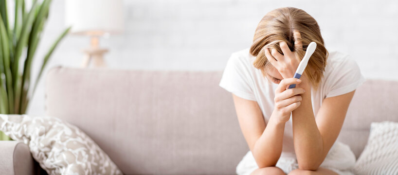 Fototapeta Lost young woman with pregnancy test sitting on couch at home and holding her head, unplanned pregnancy