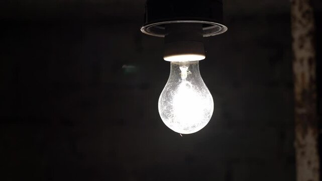 A light bulb in a dark basement blinks due to voltage drop. The basement is dark with a light bulb. Light bulb flickering in the dark.